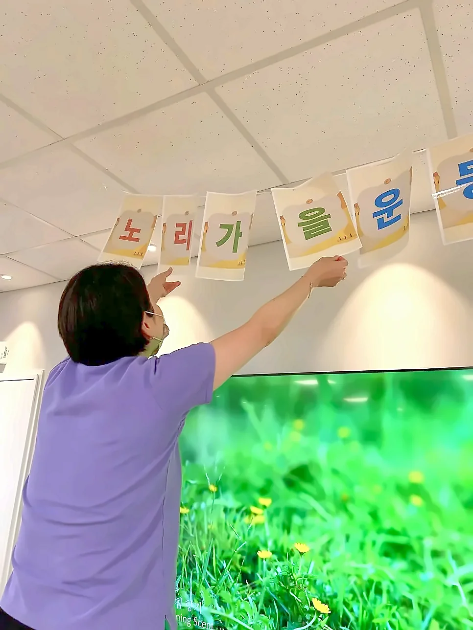 선생님이 천장 아래에 '노리 가을 운동' 글자 현수막을 거는 뒷모습