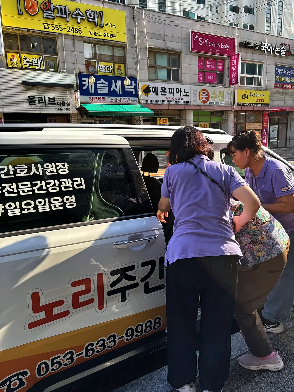 노리주간보호센터 글씨를 붙인 송영차량 옆에서 선생님 두 분이 어르신의 승차를 돕는 모습